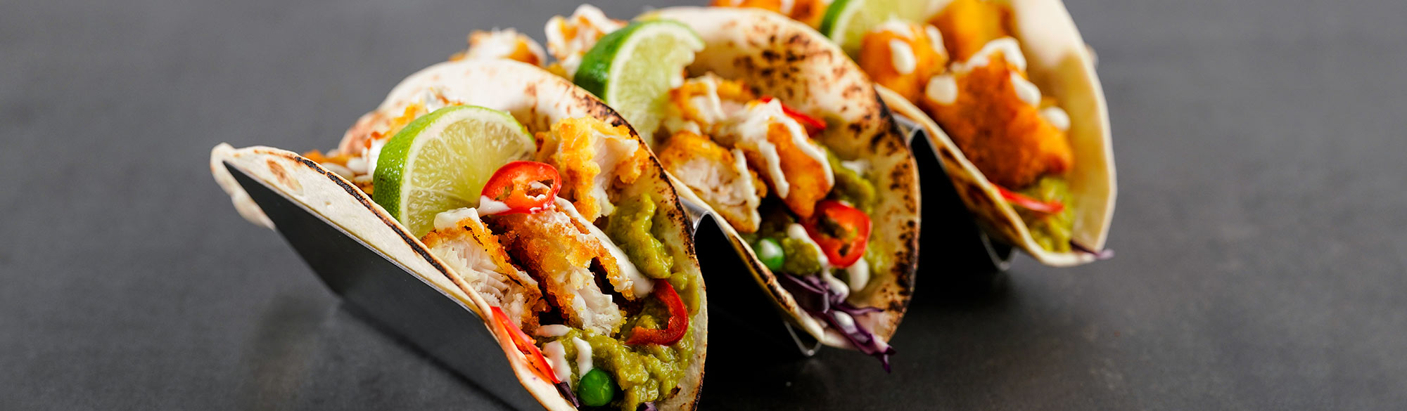 Tre fisktacos i ett serveringsställ. Frasiga fiskpinnar delade i bitar tillsammans med ärtröra, chili och lime.