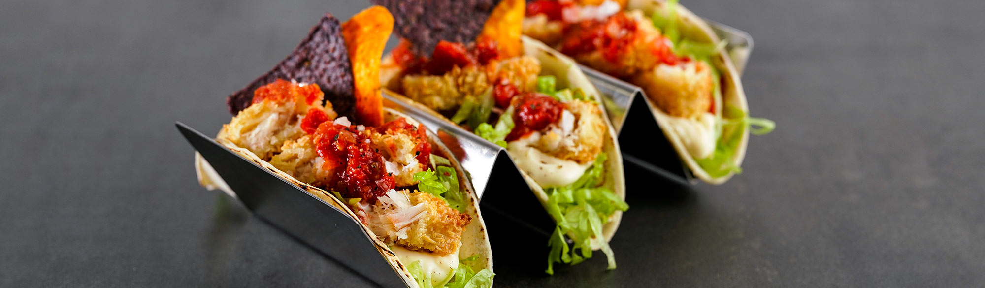 Tre fisktacos i serveringsställ. Delade fiskpinnar, sallad, salsa och nachos.