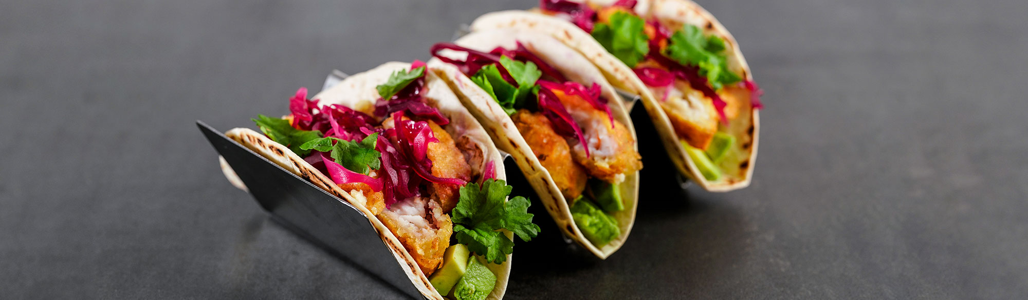 Tre fisktacos i serveringsställ. Fish n´Chips, avokadoröra, inlagdspetskål och korianderblad.